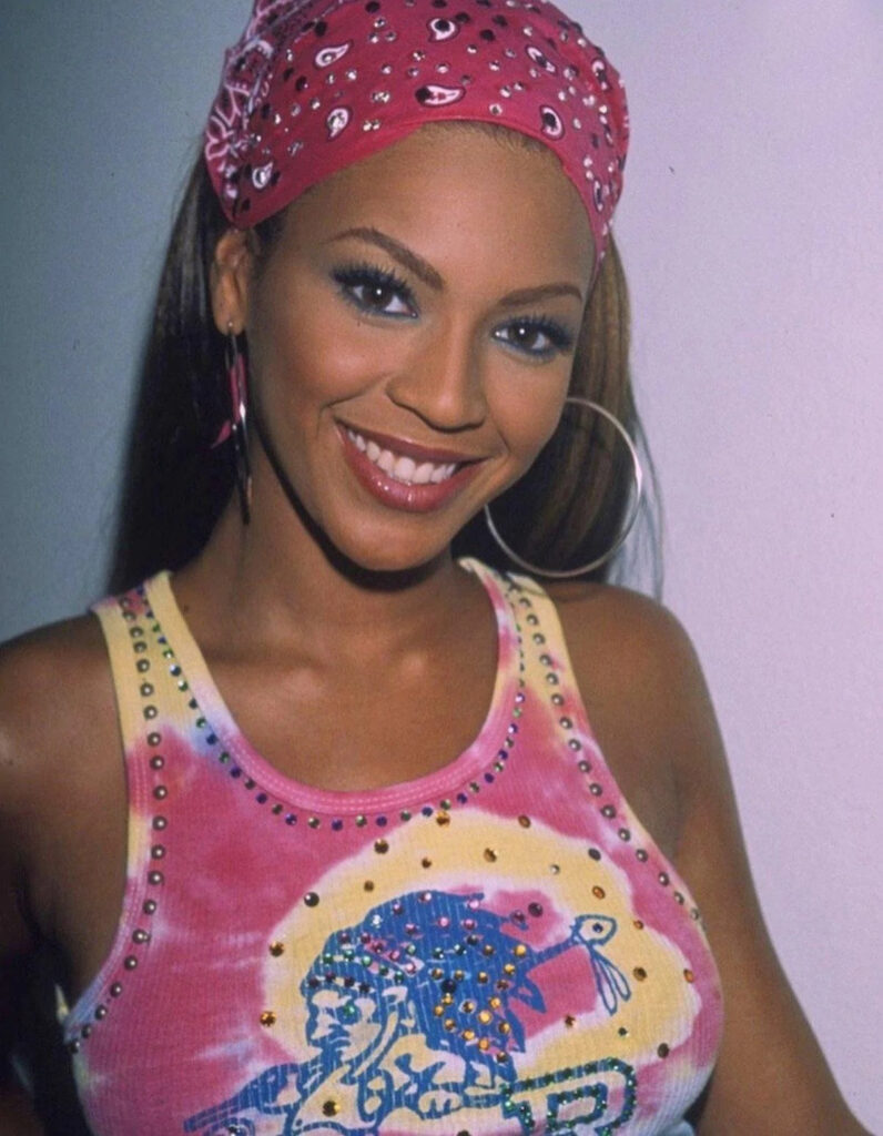 Uśmiechnięta kobieta w stylizacji Y2K – kolorowej koszulce tie-dye z grafiką i ćwiekami oraz różowej bandanie we wzory. Przykład, co to Y2K w modzie: charakterystyczne kolory, błysk i inspiracje przełomem lat 90. i 2000.