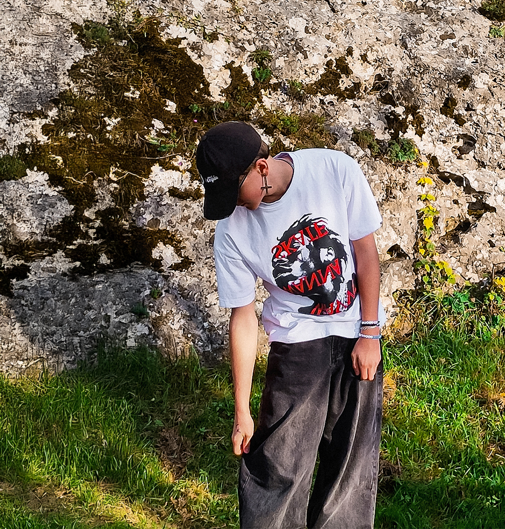 Nastolatek w czapce i białej koszulce SKATE z grafiką streetwearową stojący przy skale - styl deskorolkowy, streetwearowy