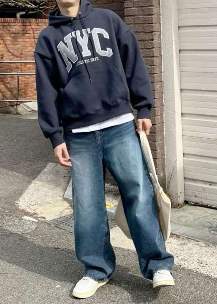 Stylizacja streetwear: granatowa bluza z kapturem NYC, szerokie jeansy, białe sneakersy i torba typu tote bag – moda uliczna 2025.