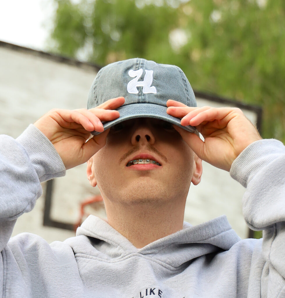 Czapka z daszkiem na modelu. Czapka z haftowanym logo H marki Highwave, w szarej bluzie, trzymający daszek – streetwearowy sty