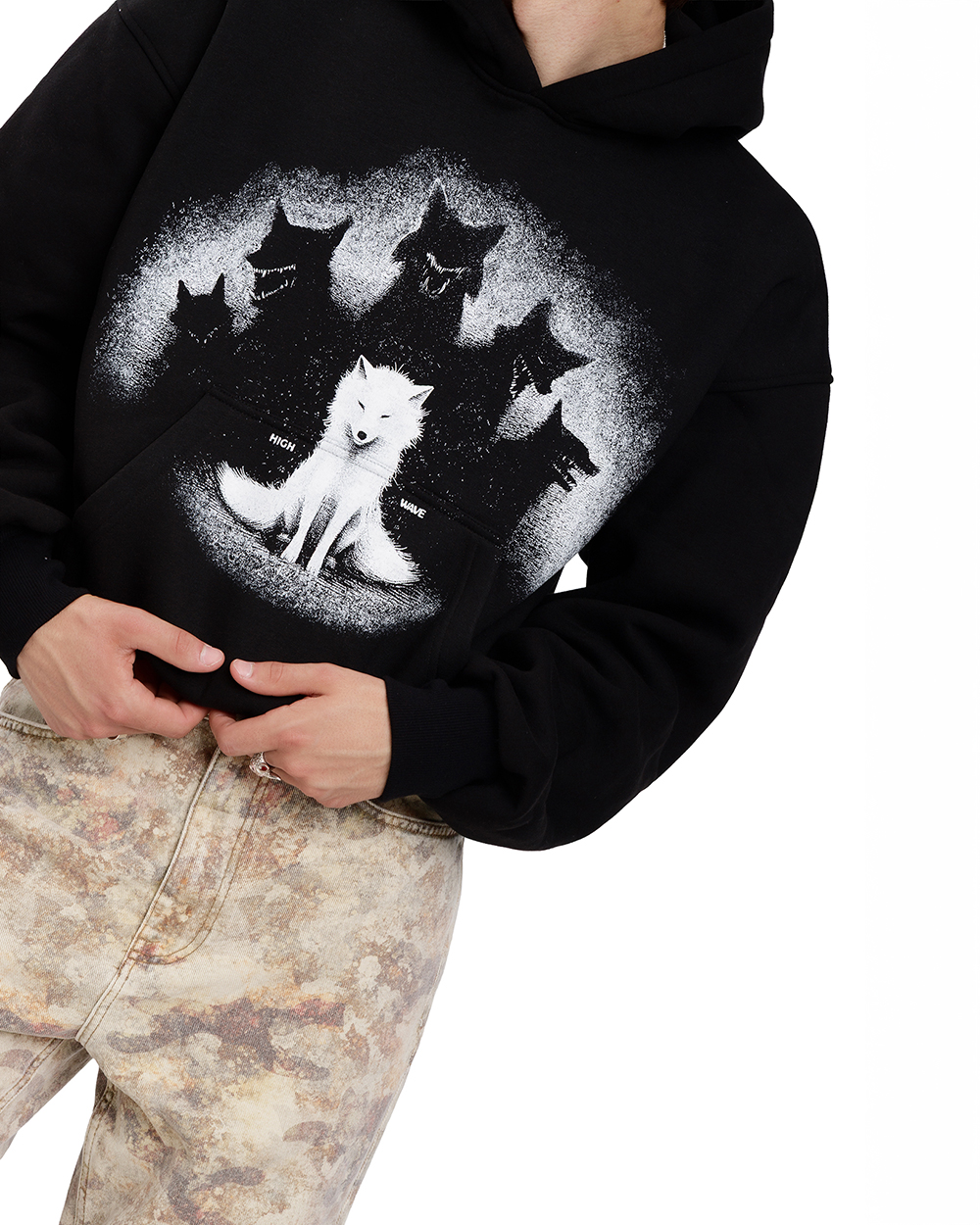 Zbliżenie na czarną bluzę z wilkiem - hoodie unisex z dużym nadrukiem białego wilka otoczonego czarnymi sylwetkami wilków. Model trzyma ręce przy kieszeni, ubrany w spodnie z marmurkowym wzorem. Streetwearowa stylizacja