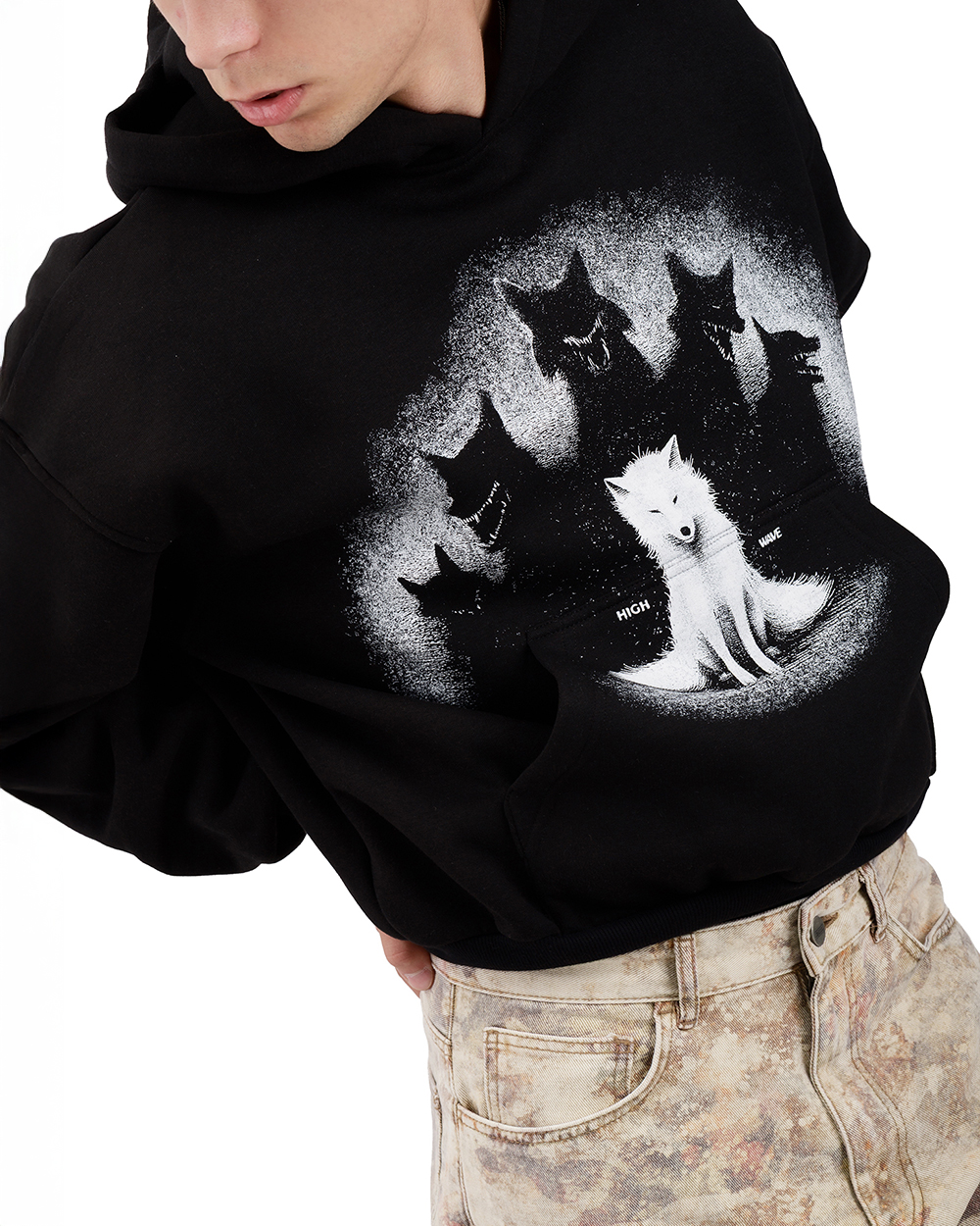 Zbliżenie na bluzę z wilkiem - czarne unisex hoodie z wyrazistym nadrukiem białego wilka i pięciu czarnych cieni wilków w tle. Model w pozie dynamicznej, ubrany w spodnie o marmurkowym wzorze. Ujęcie z boku podkreślające grafikę i detale streetwearowego stylu