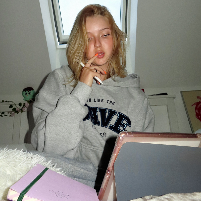 Młoda kobieta w szarej bluzie z kapturem siedzi pod oknem z tabletem i różowym notatnikiem. Trzyma długopis i wygląda na zamyśloną, czytając Newsletter Highwave