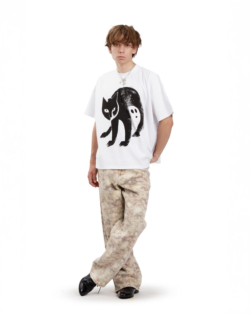 Młody mężczyzna ubrany w białą koszulkę z kotem – oversize’owy t-shirt z czarną grafiką kota i duchem w jego cieniu. Model stoi na białym tle, w jasnych marmurkowych spodniach i czarnych butach. Styl streetwear
