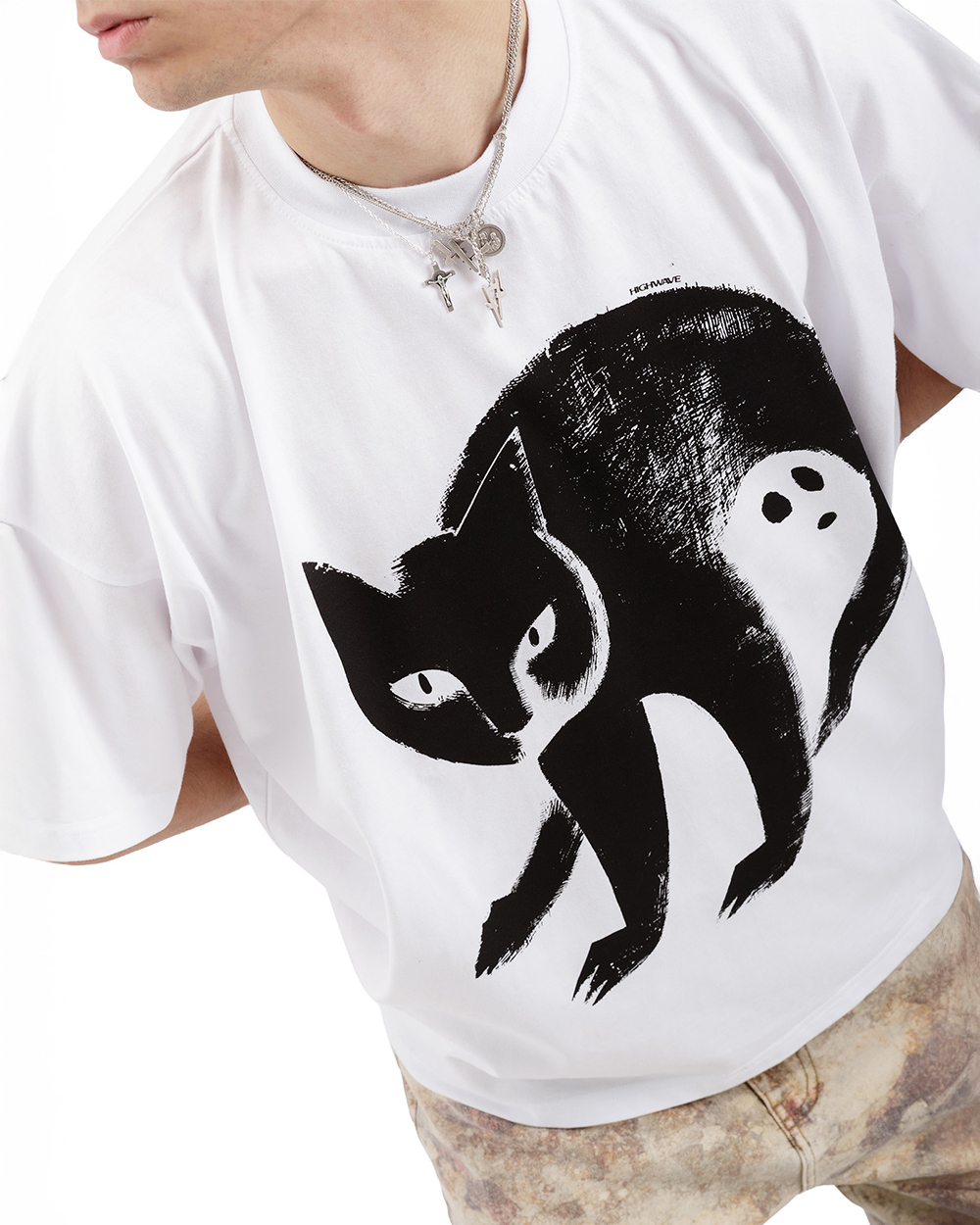 Biały t-shirt oversize z kotem – unisex koszulka 100% bawełna z czarną grafiką kota z ukrytym duchem. Model w pozie zgiętej, widoczne marmurkowe spodnie i srebrne naszyjniki. Ujęcie z góry.