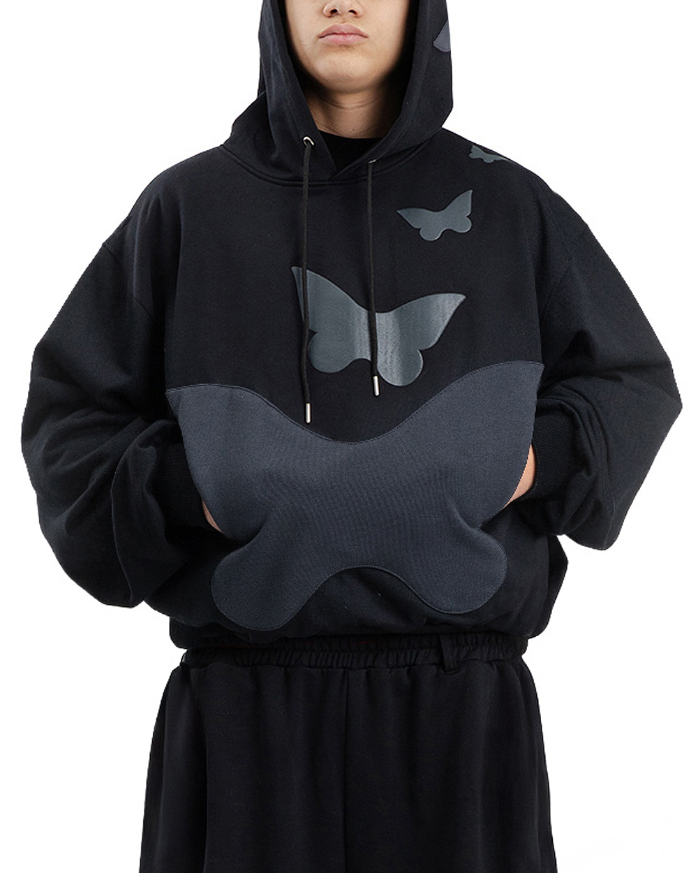 Osoba w czarnej bluzie z motylem trzyma ręce w dużej kieszeni w kształcie motyla, widoczne motylki puff print i luźny krój hoodie