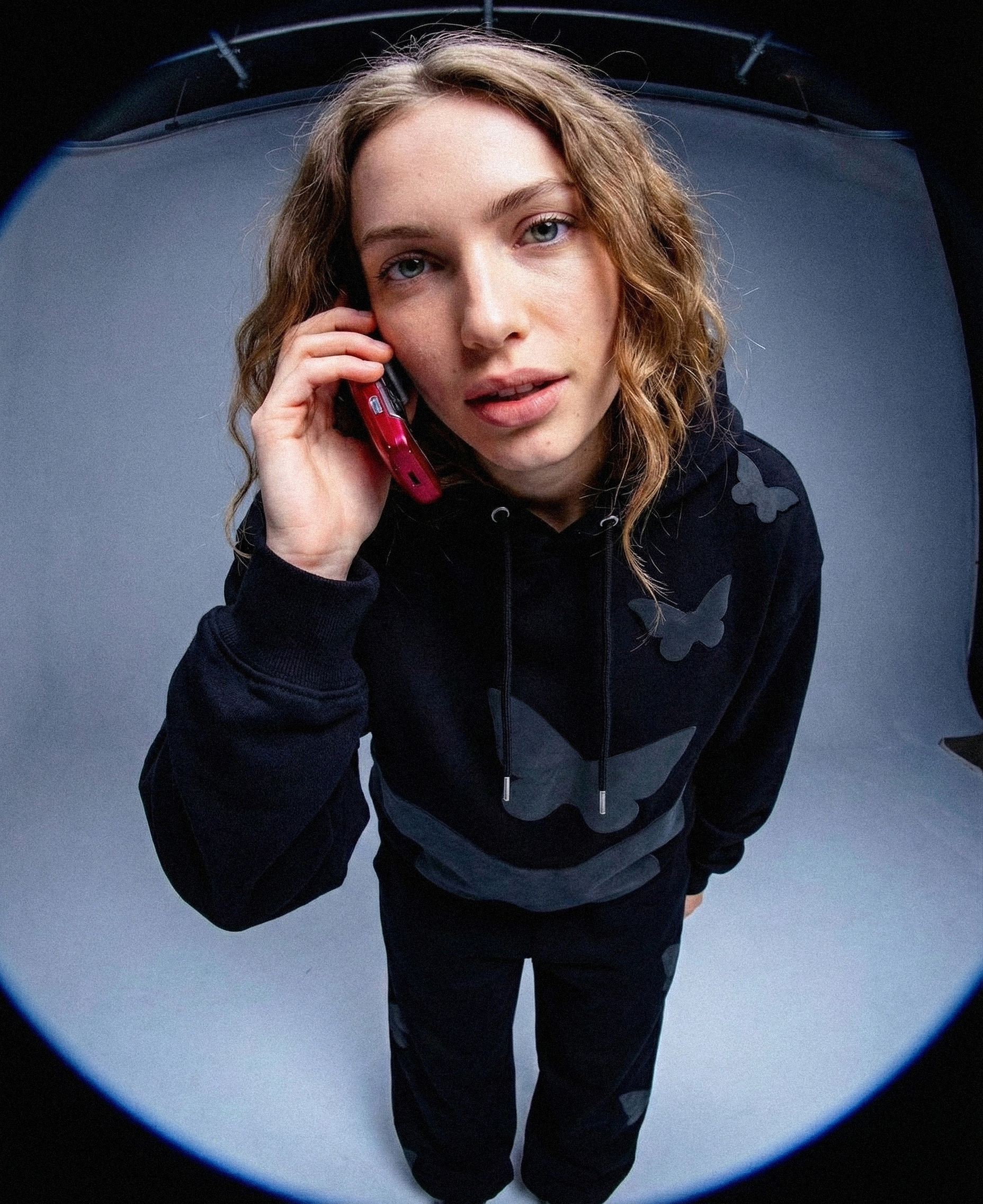 Ujęcie typu rybie oko, modelka w czarnej bluzie z motylem rozmawia przez telefon, widoczne motylki puff print i luźny krój hoodie